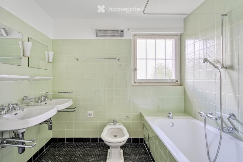 Bathroom with mint green tiles, two sinks, a bathtub, a bidet, and a mirror. Features a window and towel racks.