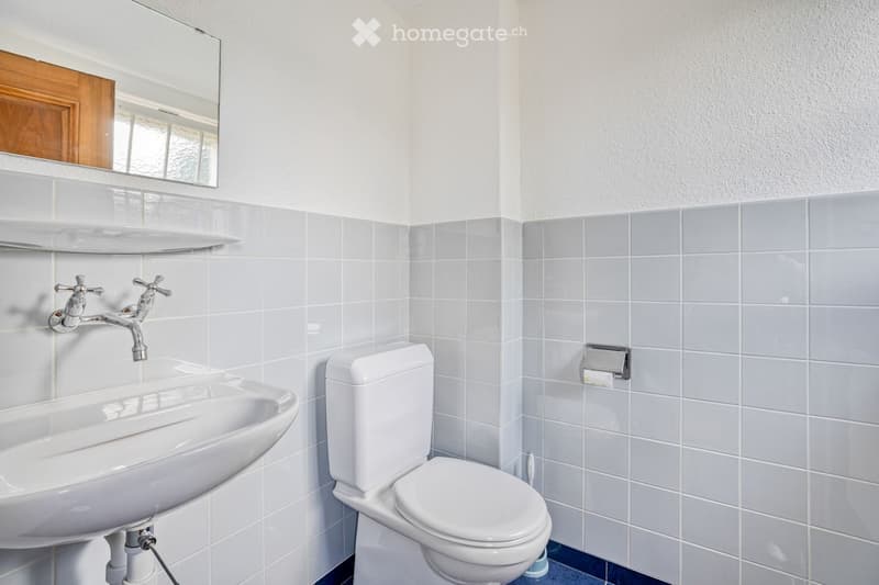 Modern bathroom with white tiles, a white sink with a chrome faucet, a toilet, a mirror with a wooden frame, and a shelf above the sink.