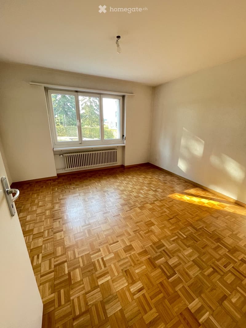 Empty room with wooden herringbone flooring, large window with view, radiator, and light fixture.