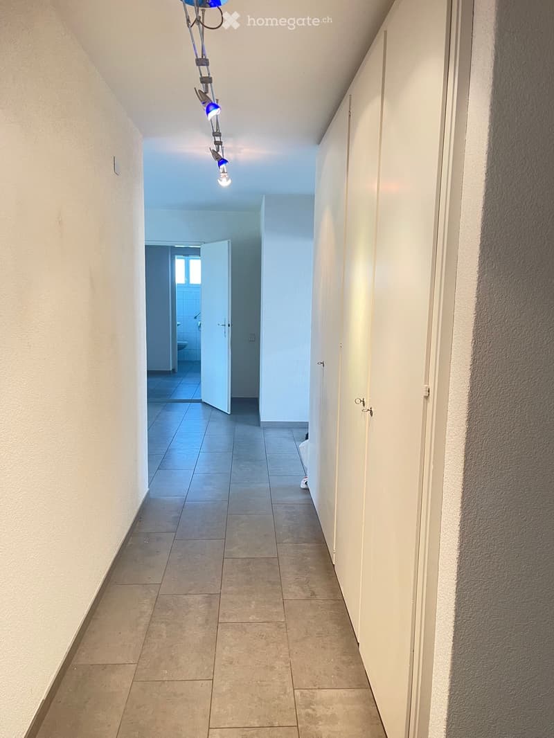 A clean, well-lit hallway with tiled flooring and white walls, featuring a partially open door leading to a bathroom and several closed doors along the right side.