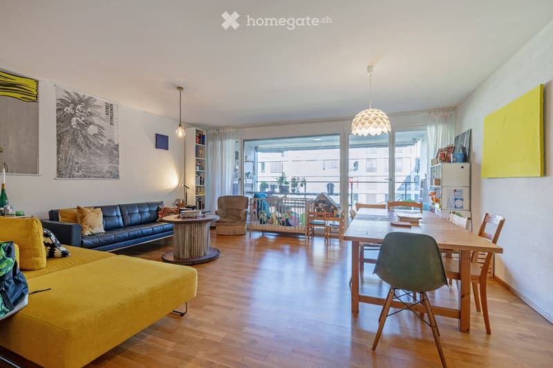 Bright, open living and dining area with wooden flooring, large windows, and a balcony. Features a yellow sofa, black leather couch, coffee table, dining table with chairs, and modern pendant light.