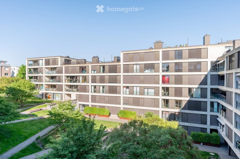 Modern multi-story residential building with balconies, large windows, and surrounding greenery.