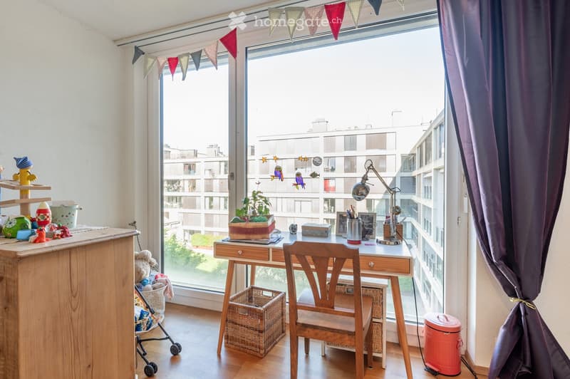 A well-lit room with a wooden desk, chair, and lamp, decorated with toys, a plant, and a curtain. A stroller and a trash bin are also visible.