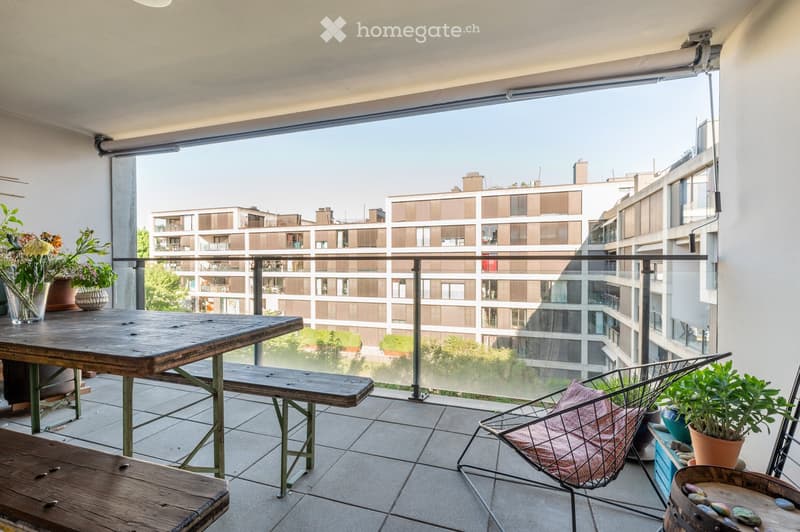 Balcony with wooden table, bench, metal chair, and potted plants, overlooking a modern building complex.