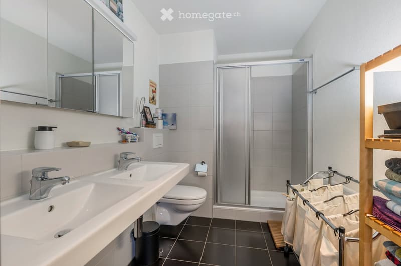 Modern bathroom with double sink, mirror, toilet, glass-enclosed shower, and wooden shelf with towels.