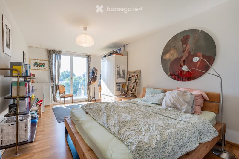 Cozy bedroom with wooden flooring, large bed with patterned bedding, a chair, a cabinet, a shelf with books, a small table, and a large round painting on the wall.