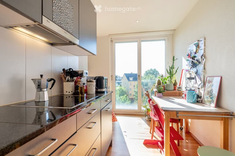 Modern kitchen with white cabinets, stainless steel appliances, induction stove, coffee maker, and a small dining area with a table and chairs. A large window offers a view of the outside, and various kitchen items and decorations are visible.