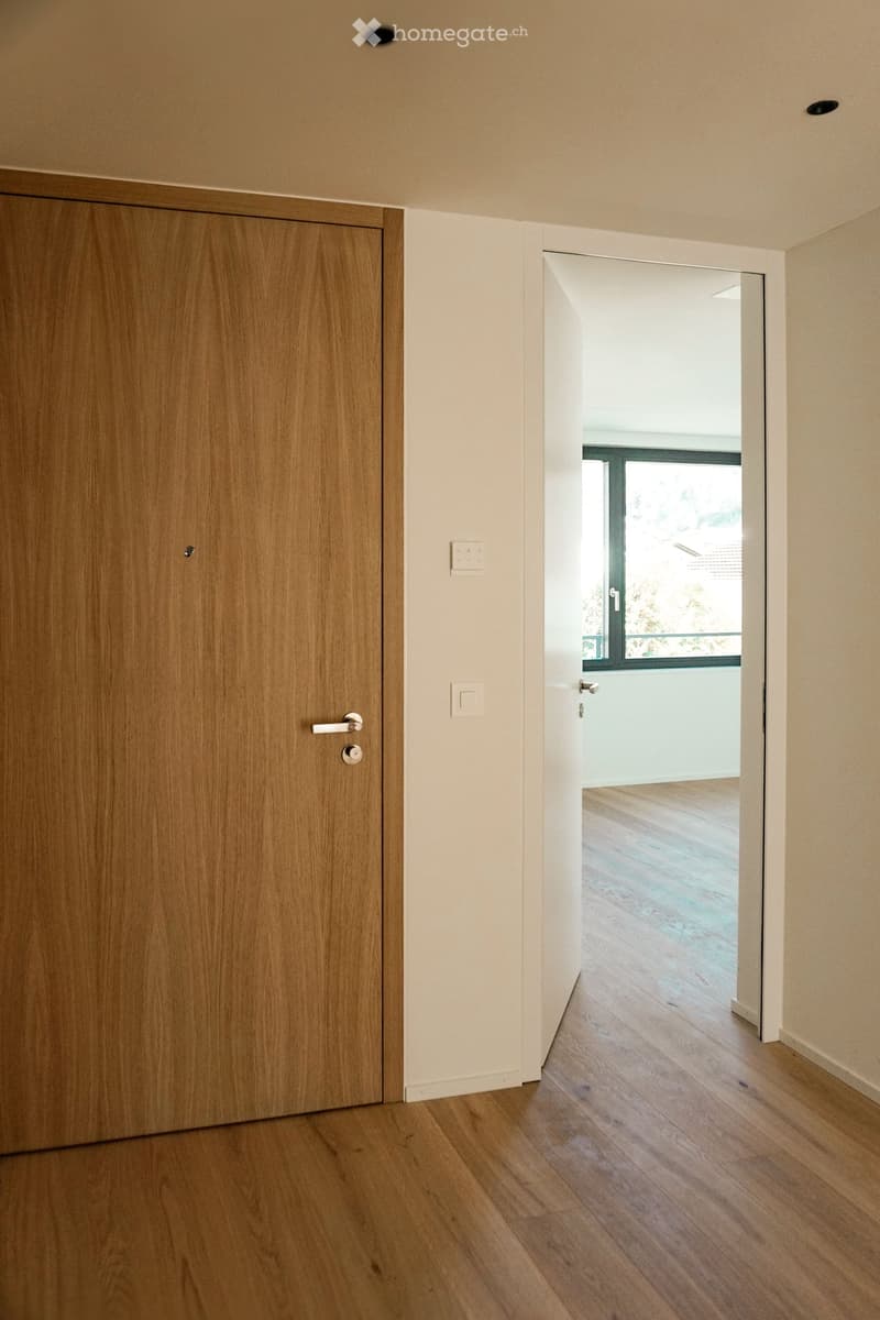 A modern hallway with light wooden flooring, a closed wooden door on the left, and an open doorway leading to a bright room with large windows and wooden flooring.