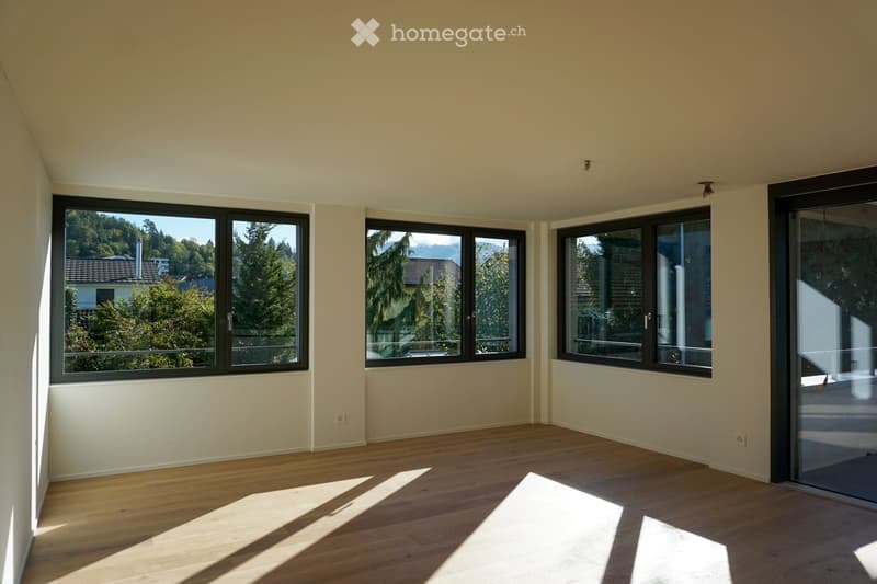 Empty room with wooden flooring and large windows offering a scenic view of trees and mountains. The space is unfurnished, with natural light streaming in.