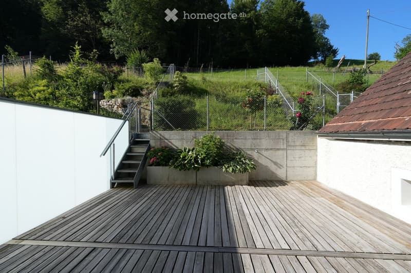 Wooden deck with metal railing, steps leading up, concrete planter with plants, and a view of a green, fenced yard.