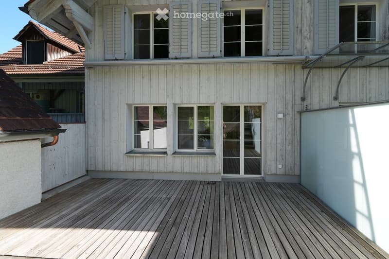 A two-story wooden house with white siding, multiple windows, and a wooden deck in front.