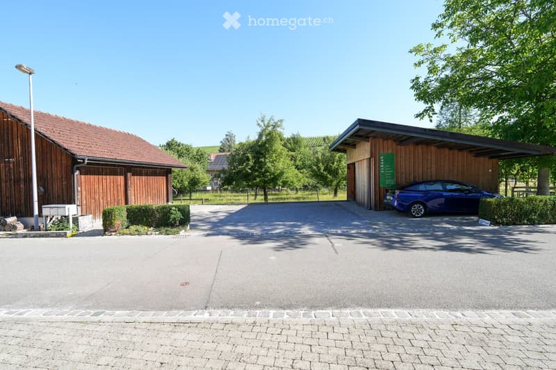 A two-story wooden house with a red-tiled roof, a covered garage with a blue car parked inside, and a well-maintained driveway surrounded by greenery.