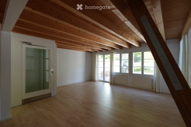 An empty room with wooden flooring and exposed wooden beams on the ceiling. Large windows and a sliding glass door lead to an outdoor area. The walls are white, and there are radiators beneath the windows.