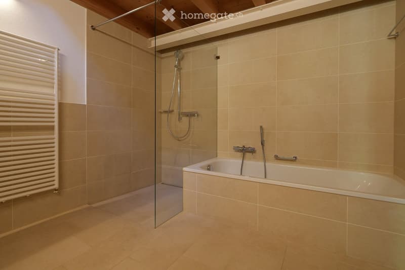 Modern bathroom with a bathtub, glass-enclosed shower, and a radiator. Beige tiles and wooden ceiling beams.