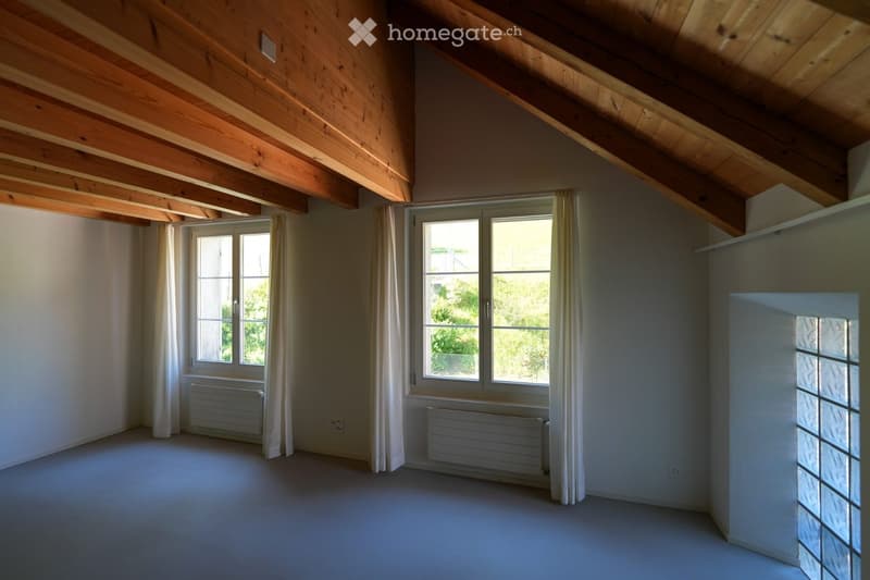 Empty attic room with wooden slatted ceiling, large windows with white curtains, radiators, and neutral-colored walls and floor.