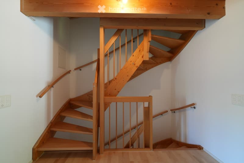 Wooden staircase with handrails and a safety gate, leading to an upper level. The walls are white, and the floor is wooden.