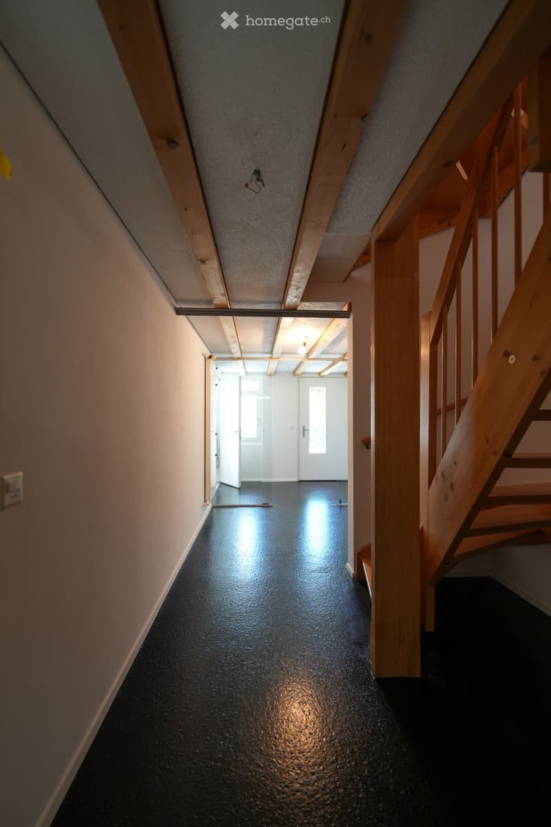 Empty hallway with wooden beams, black glossy floor, and white walls. A staircase is visible on the right side, leading upwards.