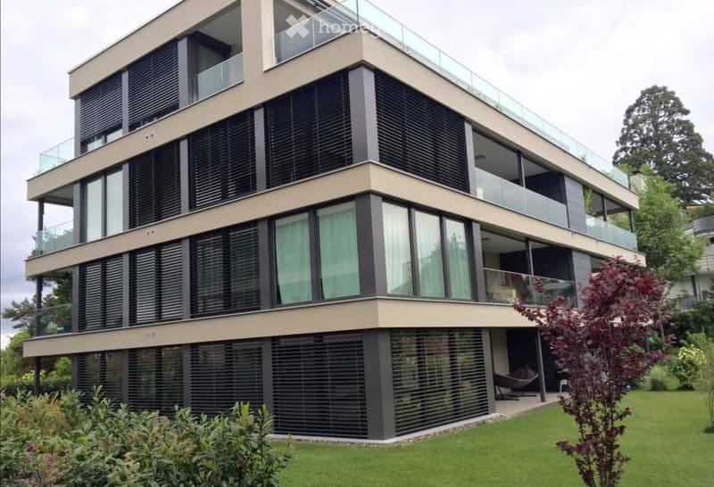 Modern multi-story building with large windows, balconies, and contemporary design, surrounded by a well-maintained lawn and garden.