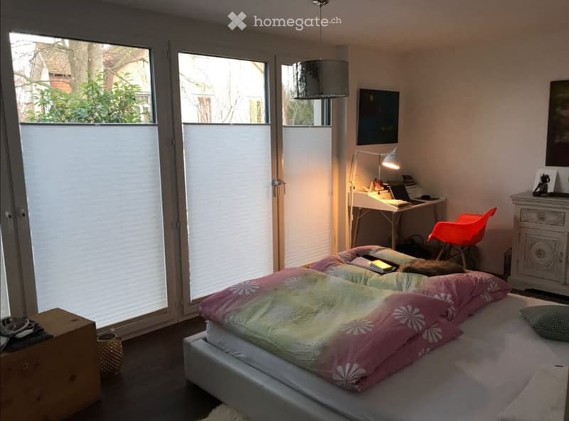 Bright bedroom with large windows, a double bed with colorful bedding, a desk with a chair, a lamp, and a cabinet.