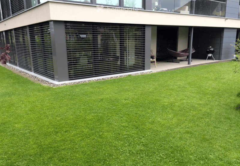 Modern house exterior with a well-maintained lawn, a covered patio area, and outdoor furniture including a hammock and grill.