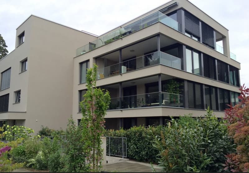 Modern multi-story residential building with large glass balconies and contemporary design, surrounded by lush greenery.