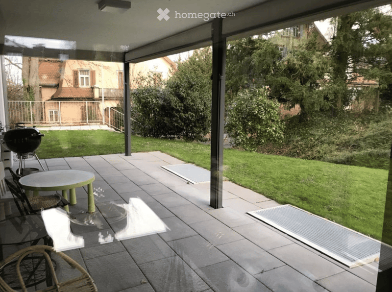 Covered patio with tiled floor, glass walls, outdoor furniture including a table, chairs, and a grill, overlooking a green lawn and neighboring houses.