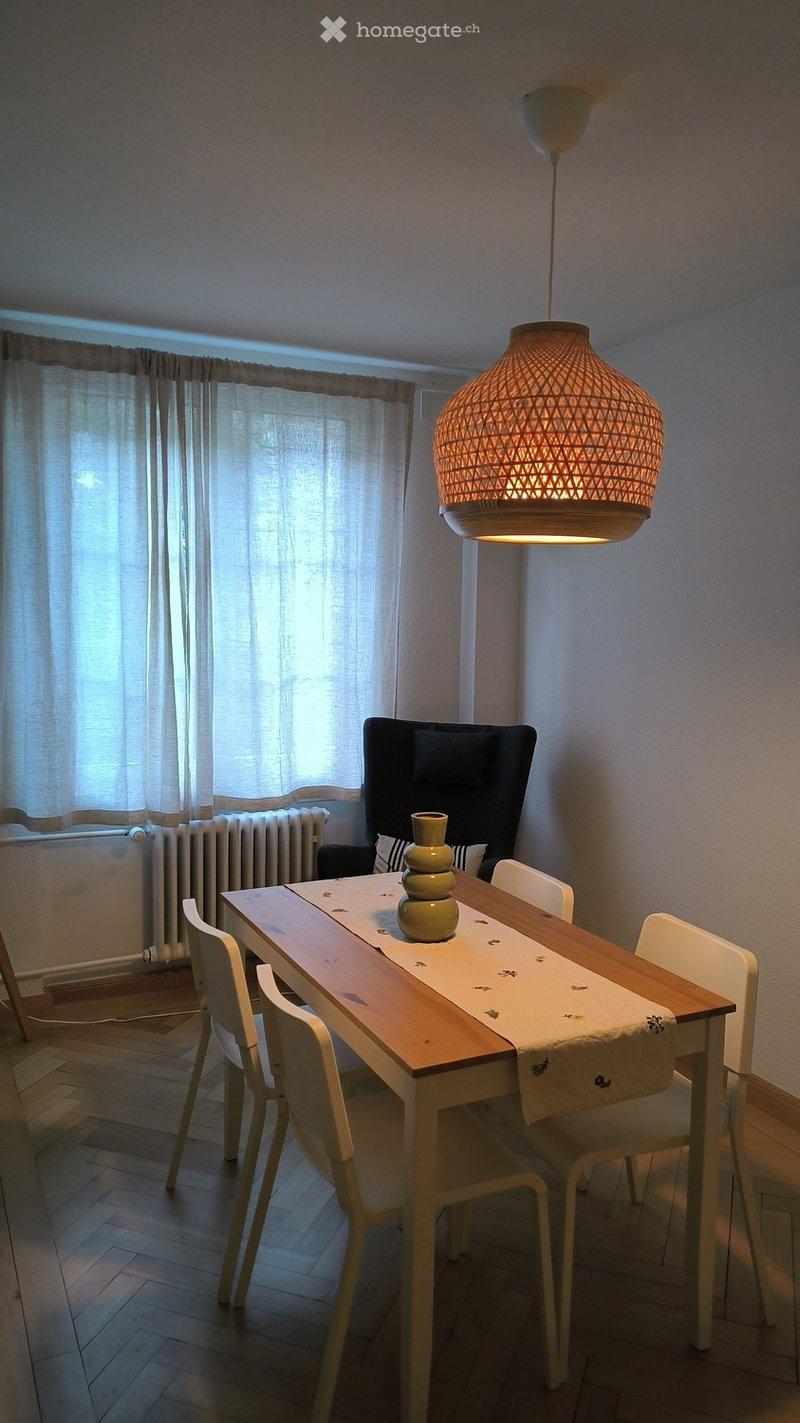 Dining area with a wooden table, white chairs, a sofa, and a decorative vase on the table. Natural light filters through sheer curtains, and a woven pendant lamp hangs from the ceiling.