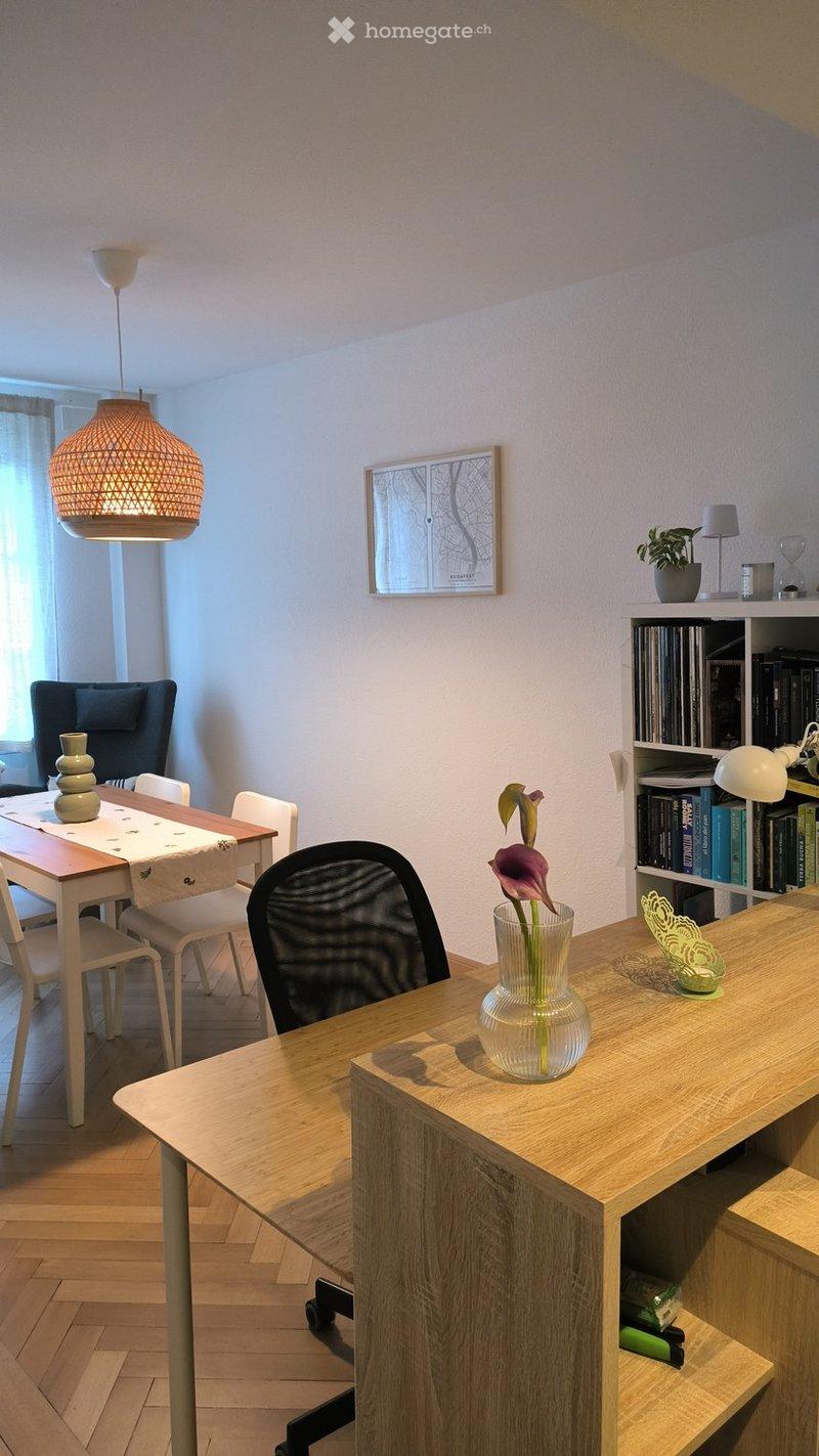 A modern, well-lit room with a wooden floor, a hanging woven lamp, a dining table with chairs, a desk with a vase of flowers, and a bookshelf filled with books and decorative items.