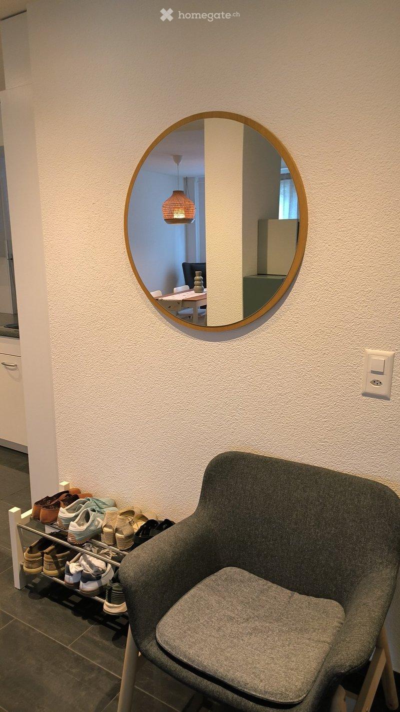 A hallway with a grey chair and a shoe rack holding multiple pairs of shoes. A round mirror on the wall reflects a dining area with a table, chairs, and a hanging lamp.