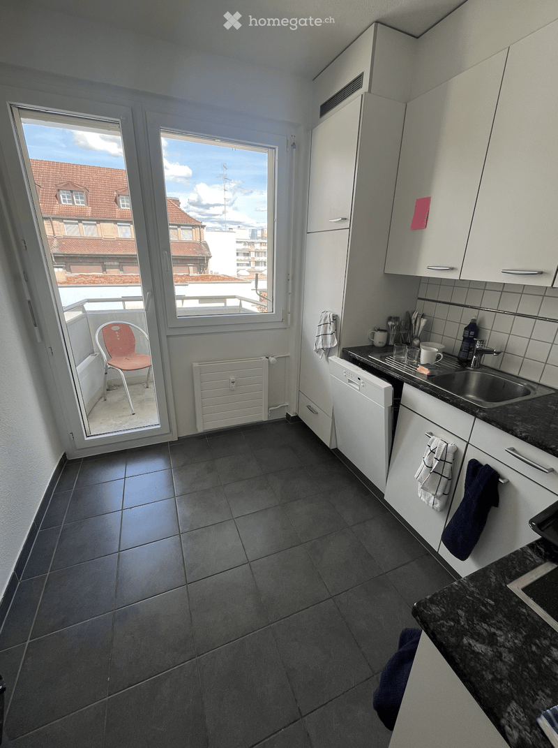 Modern kitchen with white cabinets, dark tiled floor, and a view of the city through large windows. Includes a red chair on the balcony, a dishwasher, sink, and various kitchen items.