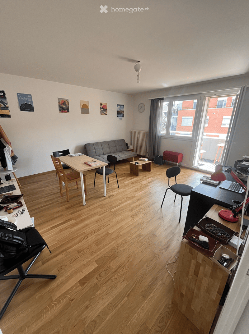 A modern, well-lit living space with wooden flooring, a grey sofa, a wooden dining table with chairs, a desk with a laptop, and various decorative items including artwork and a clock.