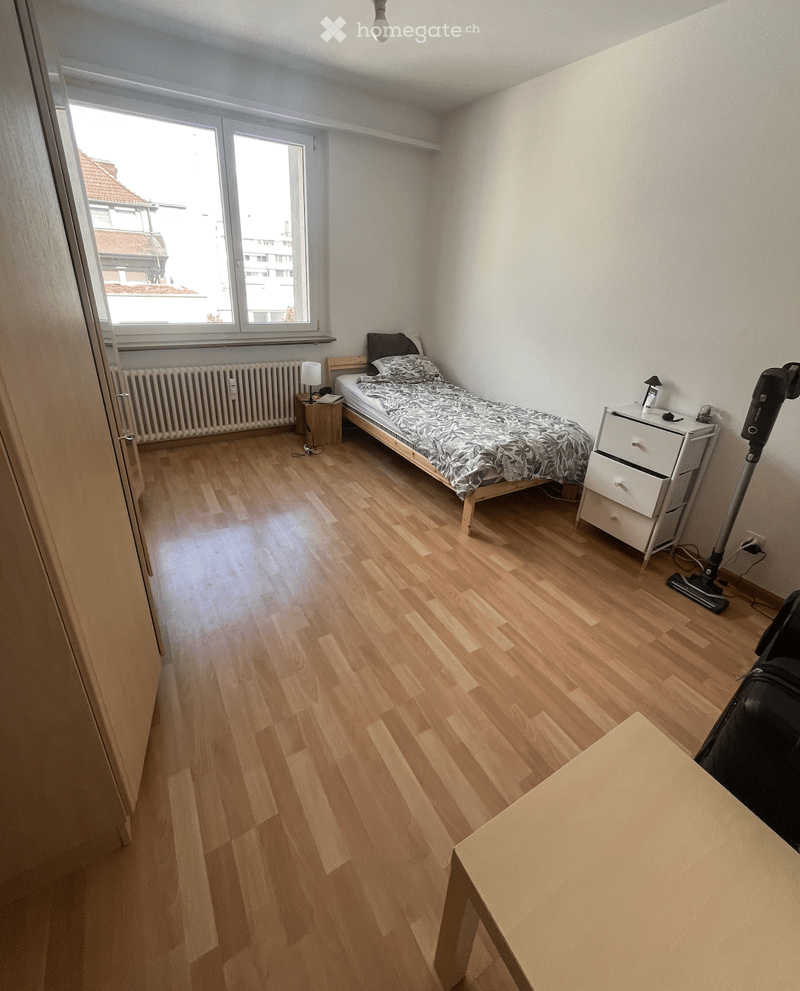 Cozy bedroom with wooden flooring, a single bed with patterned bedding, a nightstand with a lamp, and a radiator under the window.