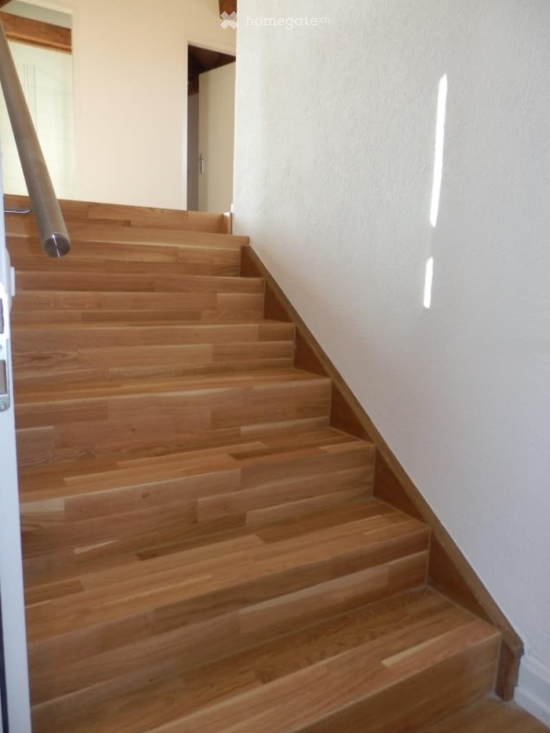 Wooden staircase with a handrail, leading up to an open doorway, with white walls and a slanted ceiling.
