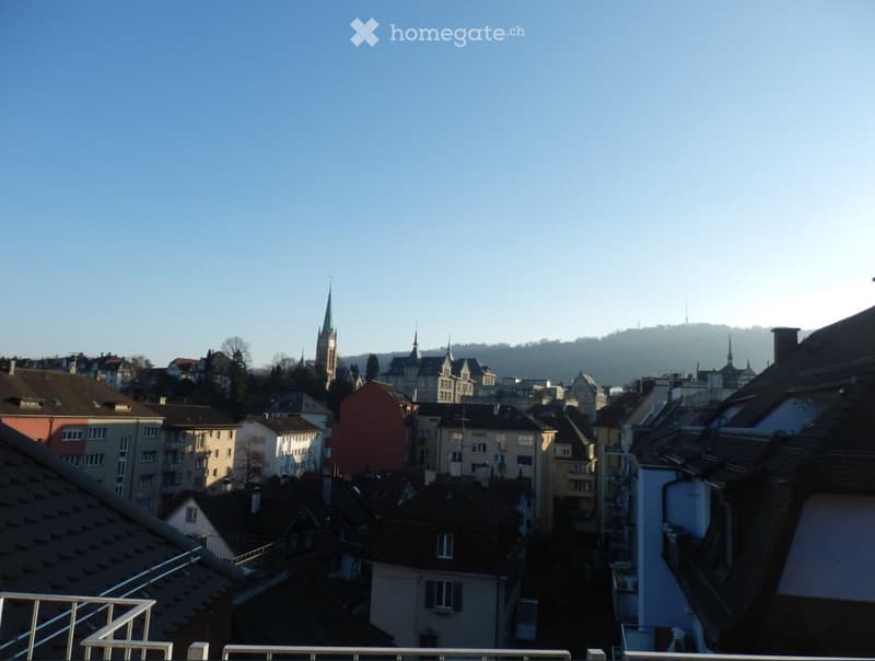 View from a balcony overlooking a cityscape with various buildings, including a prominent church tower, under a clear blue sky.