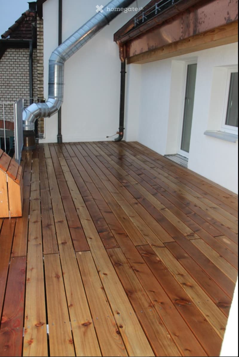 Wooden deck with a metal railing, white walls, and a metal pipe running along the side.