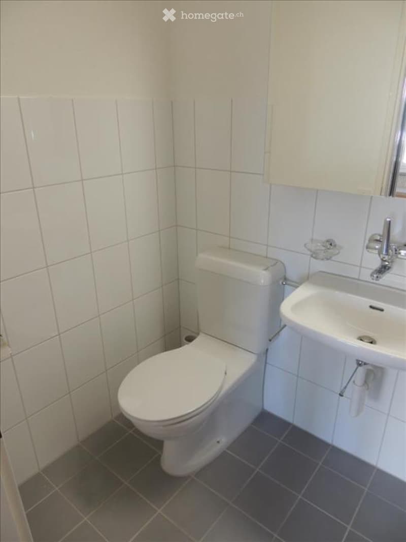 A small, clean bathroom with white tiled walls, a toilet, a sink with a mirror, and a white cabinet above the sink.