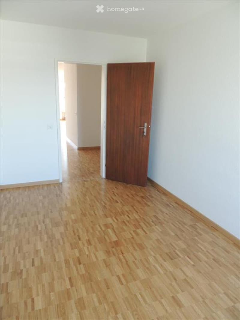 Empty room with light-colored wooden flooring and white walls. A wooden door is visible on the right side, and an open doorway leads to another room in the background.
