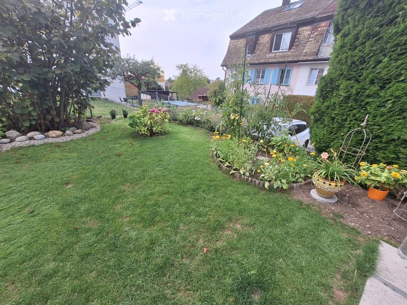 Well-maintained garden with a lawn, flower beds, potted plants, and a white car parked near the house.