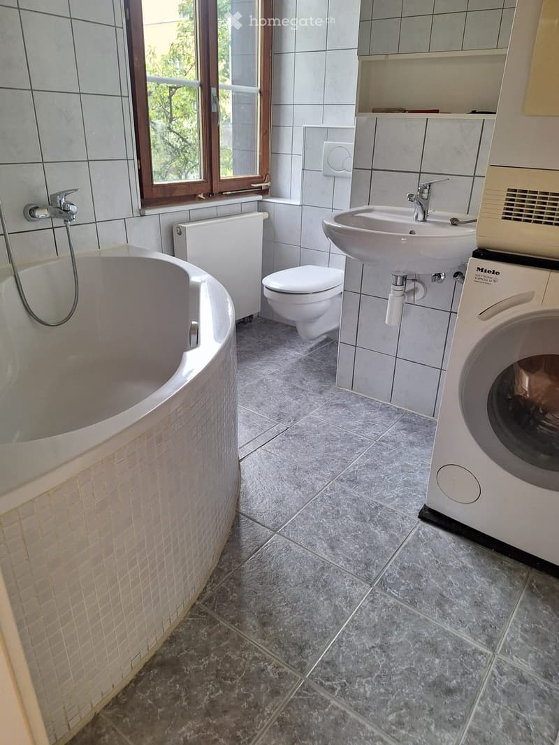 Modern bathroom with a bathtub, toilet, sink, and washing machine. Features tiled walls, a window, and a radiator.