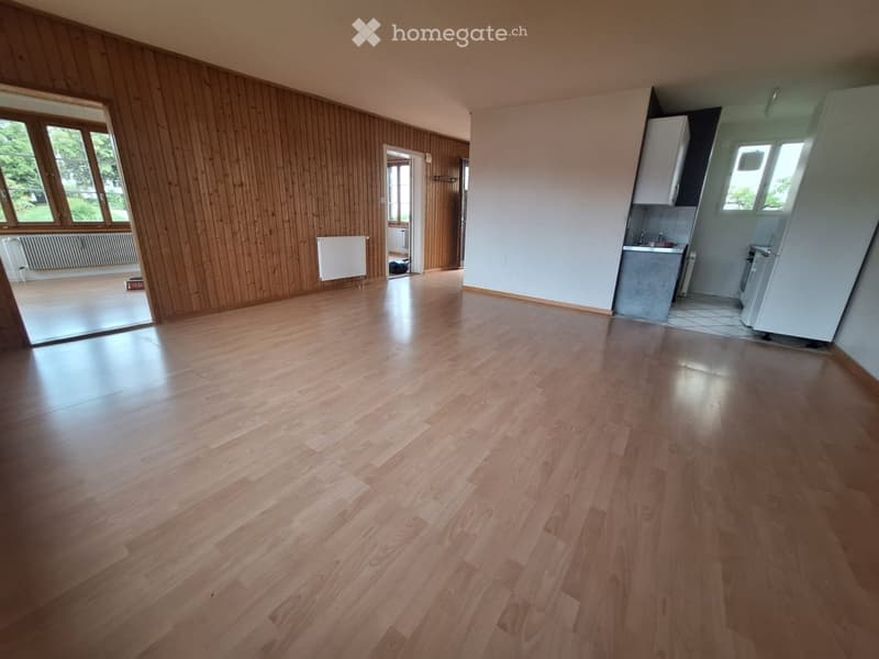 Empty room with wooden flooring, white walls, and large windows. Adjacent kitchen area with cabinets and appliances visible.