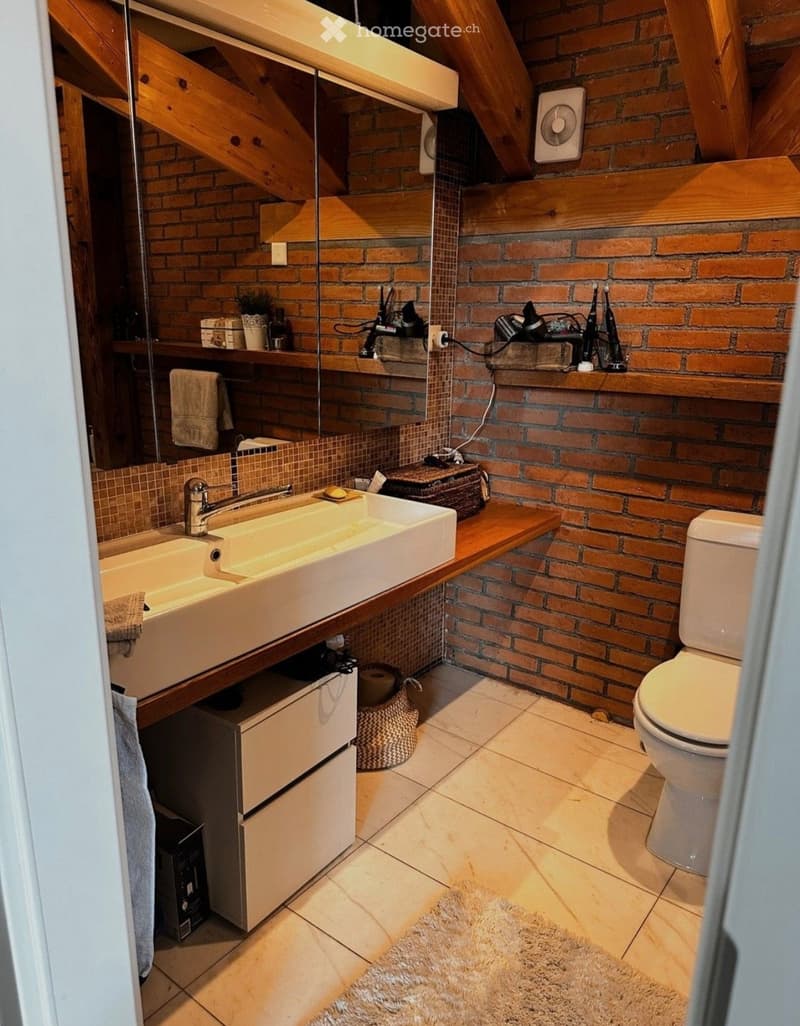 Modern bathroom with exposed brick walls, double sink vanity, toilet, mirror, and wooden shelves holding various items.