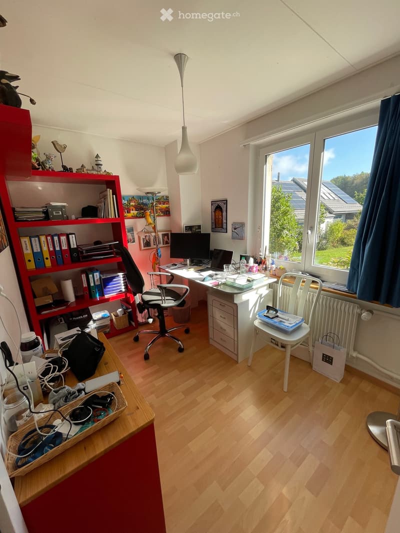 A well-lit study or office space with wooden flooring, a large window with blue curtains, a red bookshelf, a white desk, a black office chair, and various office supplies.