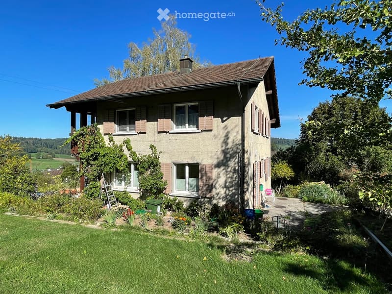 Two-story house with a tiled roof, multiple windows, and a well-maintained garden with greenery and a small patio area.