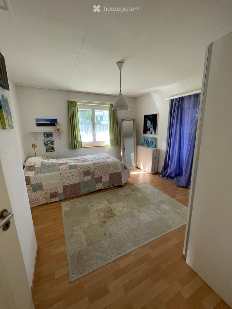 Bright bedroom with wooden flooring, large window with green and blue curtains, bed with patchwork quilt, mirror, table lamp, shelf with picture frames, and a decorative rug.