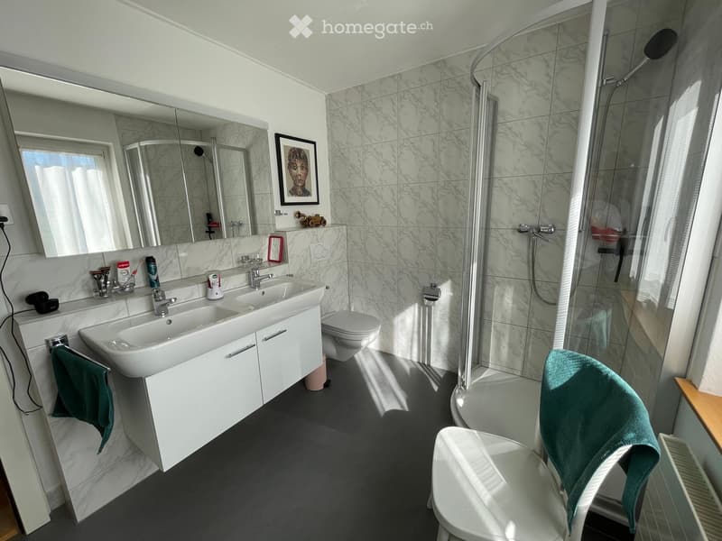 Modern bathroom with double sink, large mirror, toilet, glass-enclosed shower, and a white chair with a green towel draped over it.