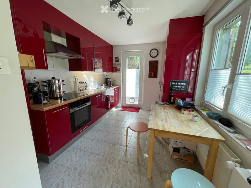 Modern kitchen with red cabinets, stainless steel appliances, a central island with a wooden table and stools, and large windows letting in natural light.