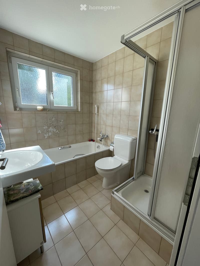 Bathroom with beige tiled walls and floor, featuring a bathtub, toilet, sink, and a glass-enclosed shower stall. A window with frosted glass is above the bathtub, and a small cabinet holds a magazine.