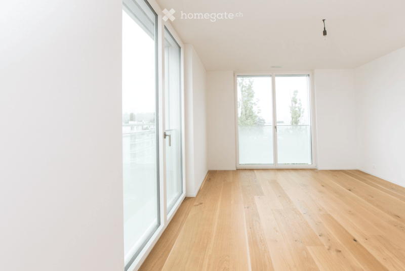 Empty room with light wooden flooring, white walls, and large glass windows and doors leading to a balcony or outdoor area.