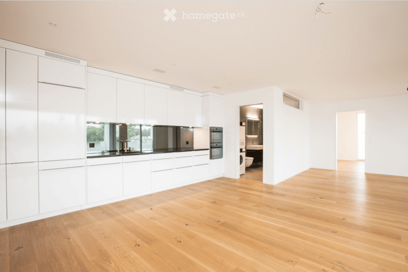 Open-plan space with light wooden flooring, white cabinetry, and a modern kitchen area visible in the background. The room is completely empty with no furniture or personal items.