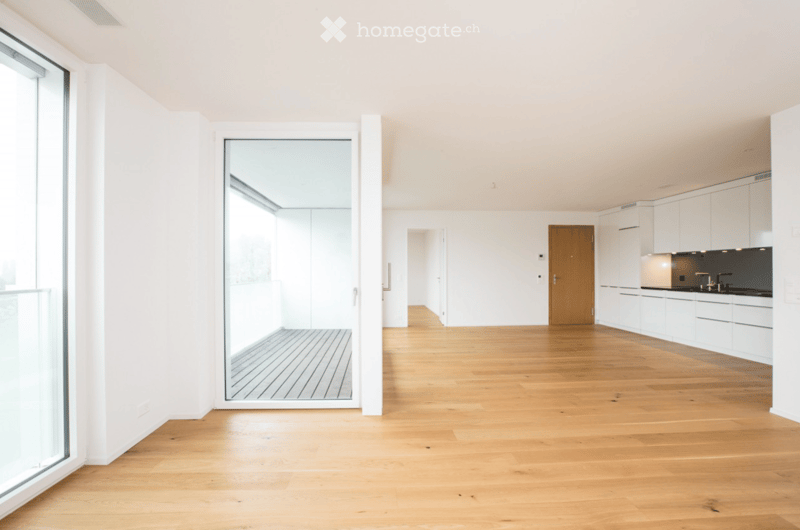 Bright, open-plan space with light wooden flooring, white walls, and large glass windows. The area includes a kitchen with white cabinets and a dark countertop, and a wooden door leading to another room.